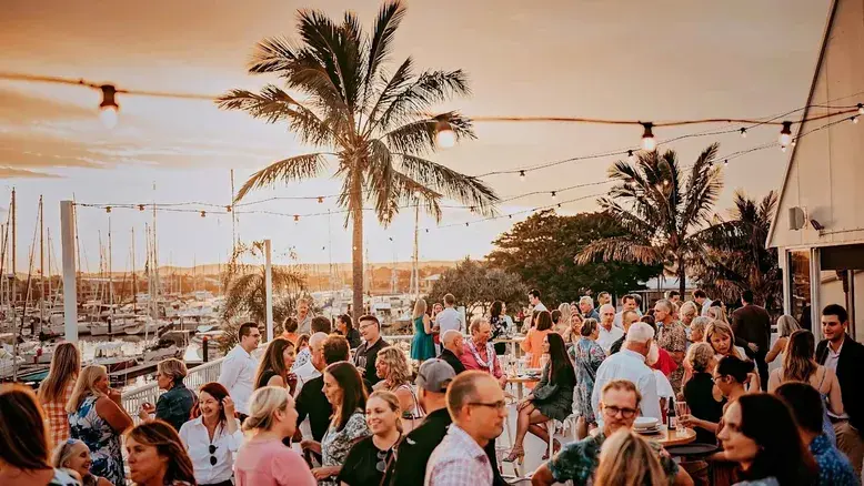 new years eve sunshine coast marina drinks at sunset with a lively crowd under festoon lights, palm trees and yachts in the harbour